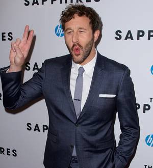 Chris O'Dowd, with a neutral expression, sits at a desk on a TV set