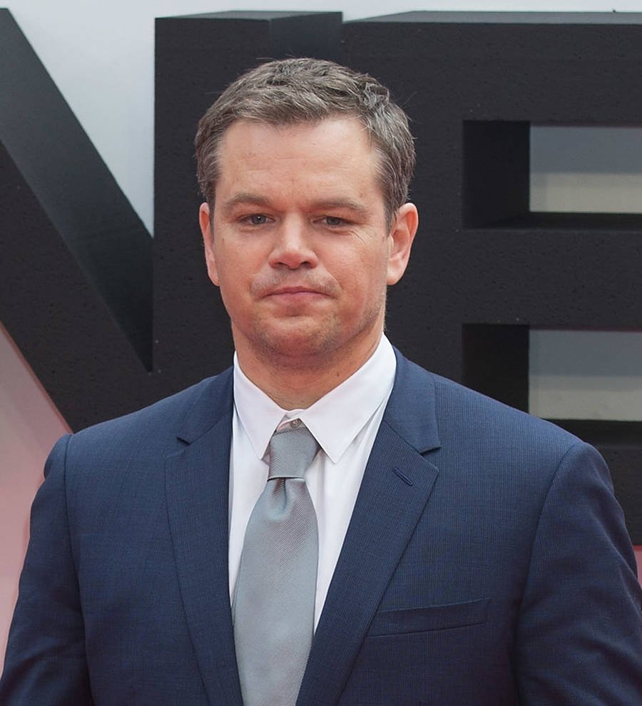 Matt Damon in a dark jacket, smiling at an event, with people in the background