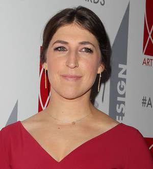 Mayim Bialik, with a sad expression, wears a black top and glasses, mourning the loss of her father