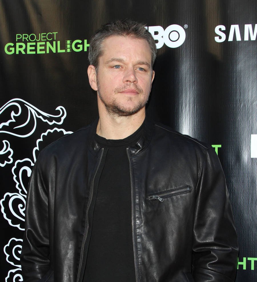 Matt Damon stands in a striped shirt, arm raised, speaking at a microphone at a press conference