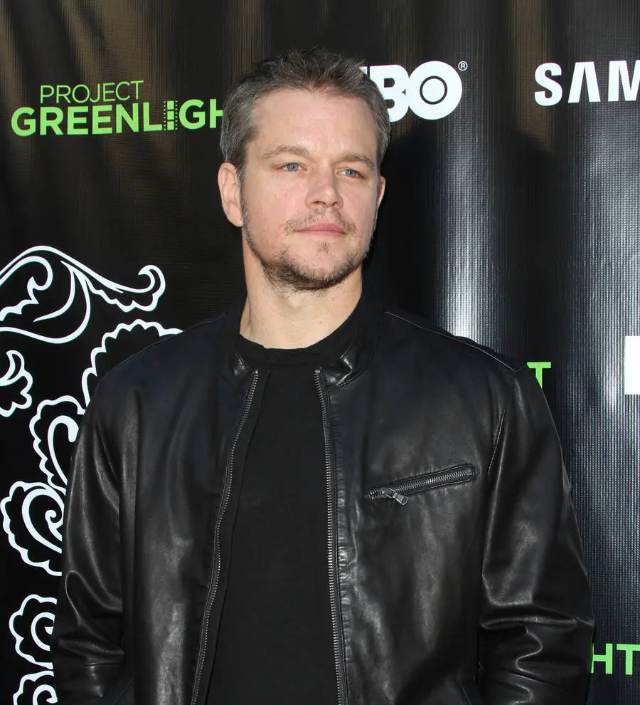 Matt Damon stands in a striped shirt, arm raised, speaking at a microphone at a press conference