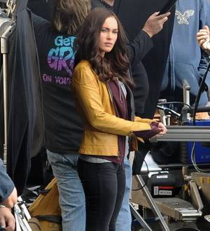 Megan Fox, in a black top and jeans, walks down a city street with dark sunglasses and a serious expression