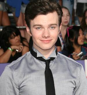 Chris Colfer beams in a blue shirt and dark blazer, standing outdoors with a green plant background