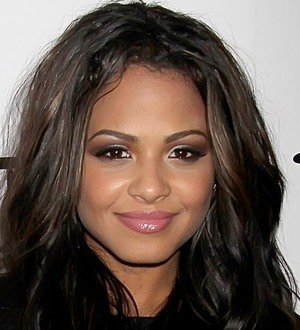 Christina Milian in a pink top poses on a beach with ocean and palm trees behind her