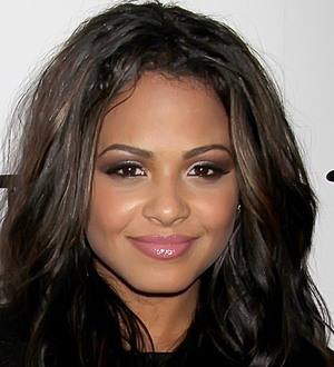 Christina Milian in a pink top poses on a beach with ocean and palm trees behind her