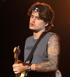 John Mayer in a dark suit, white shirt, and black tie looks at the camera. He is smiling faintly
