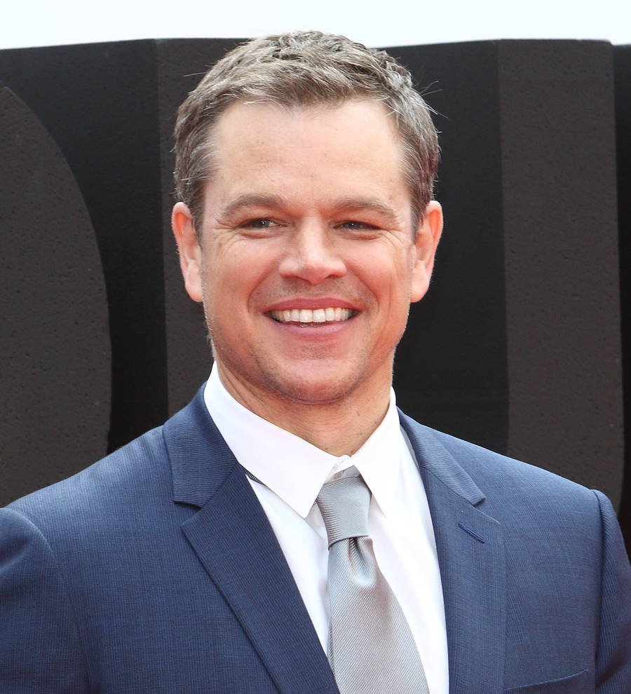 Matt Damon in a black shirt, smiling slightly with his arms crossed over a brown patterned background