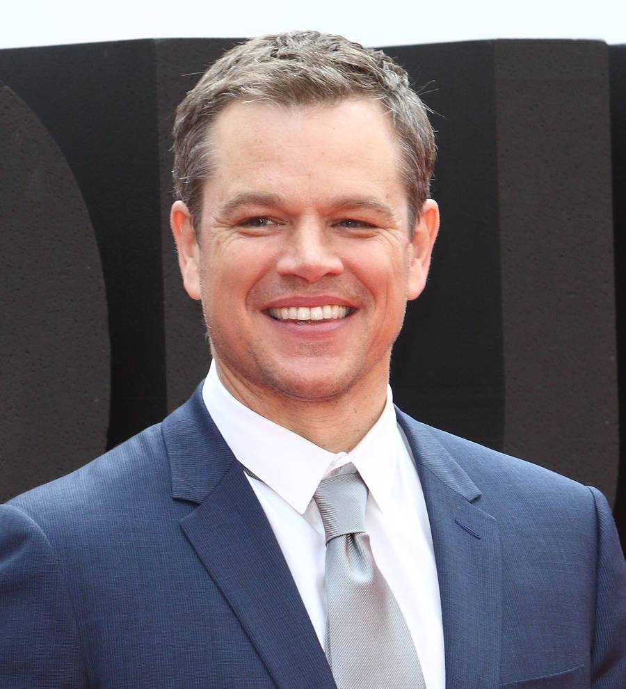 Matt Damon in a black shirt, smiling slightly with his arms crossed over a brown patterned background