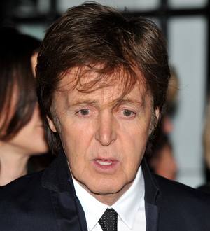 Paul McCartney smiles, gesturing with one hand, a microphone in front of him