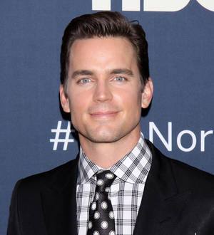 Matt Bomer with short dark hair, wearing a black shirt and black jacket, smiles in front of a red background