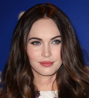 Megan Fox poses wearing a shiny gold dress, smiling and looking to the side
