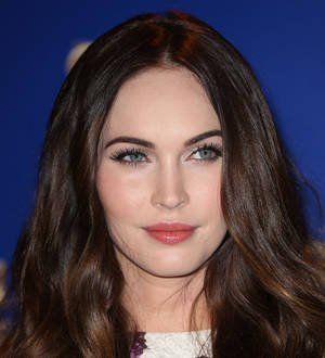 Megan Fox poses wearing a shiny gold dress, smiling and looking to the side