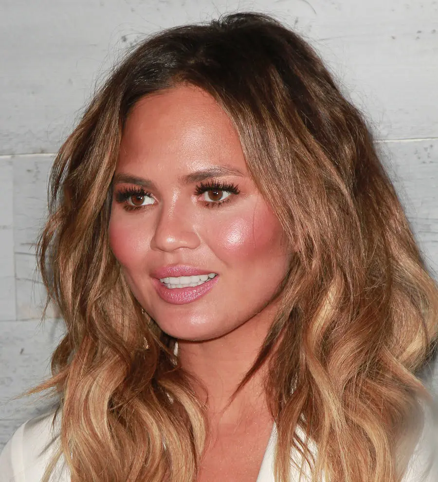 Chrissy Teigen in a red gown with a microphone, appearing to speak at an event