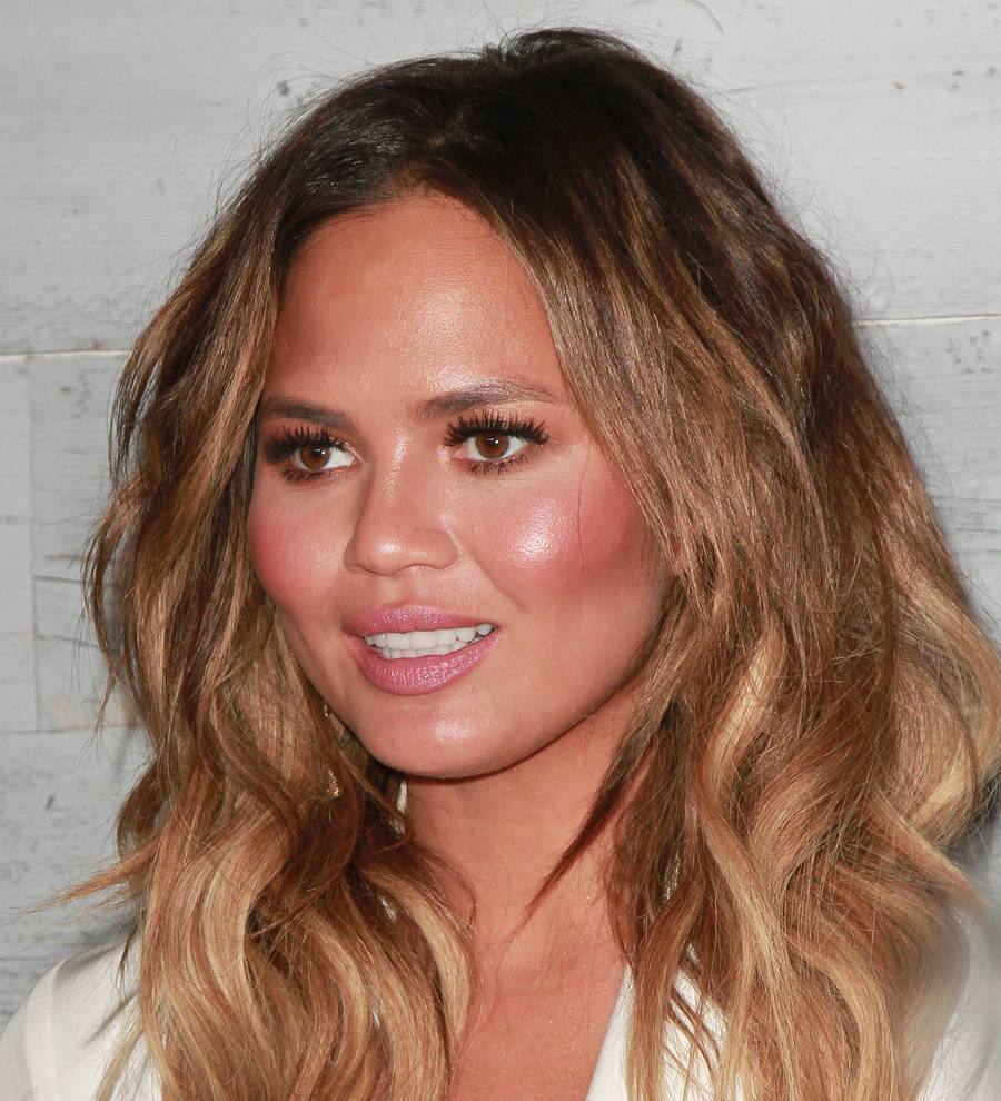 Chrissy Teigen in a red gown with a microphone, appearing to speak at an event