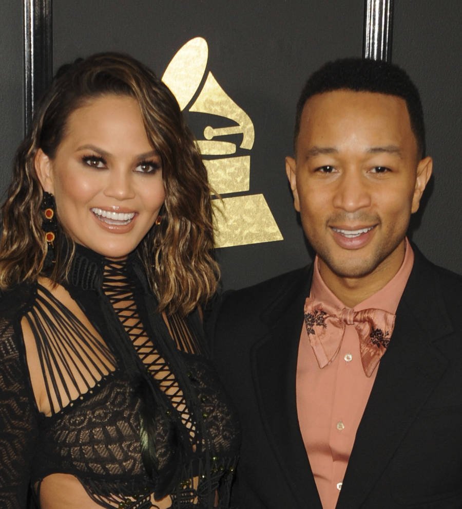 Chrissy Teigen in a red bra, black shorts, and a short sheer black robe, singing on a bed