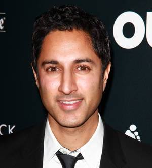 Maulik Pancholy smiles wearing a black suit jacket and white collared shirt in a close-up studio shot