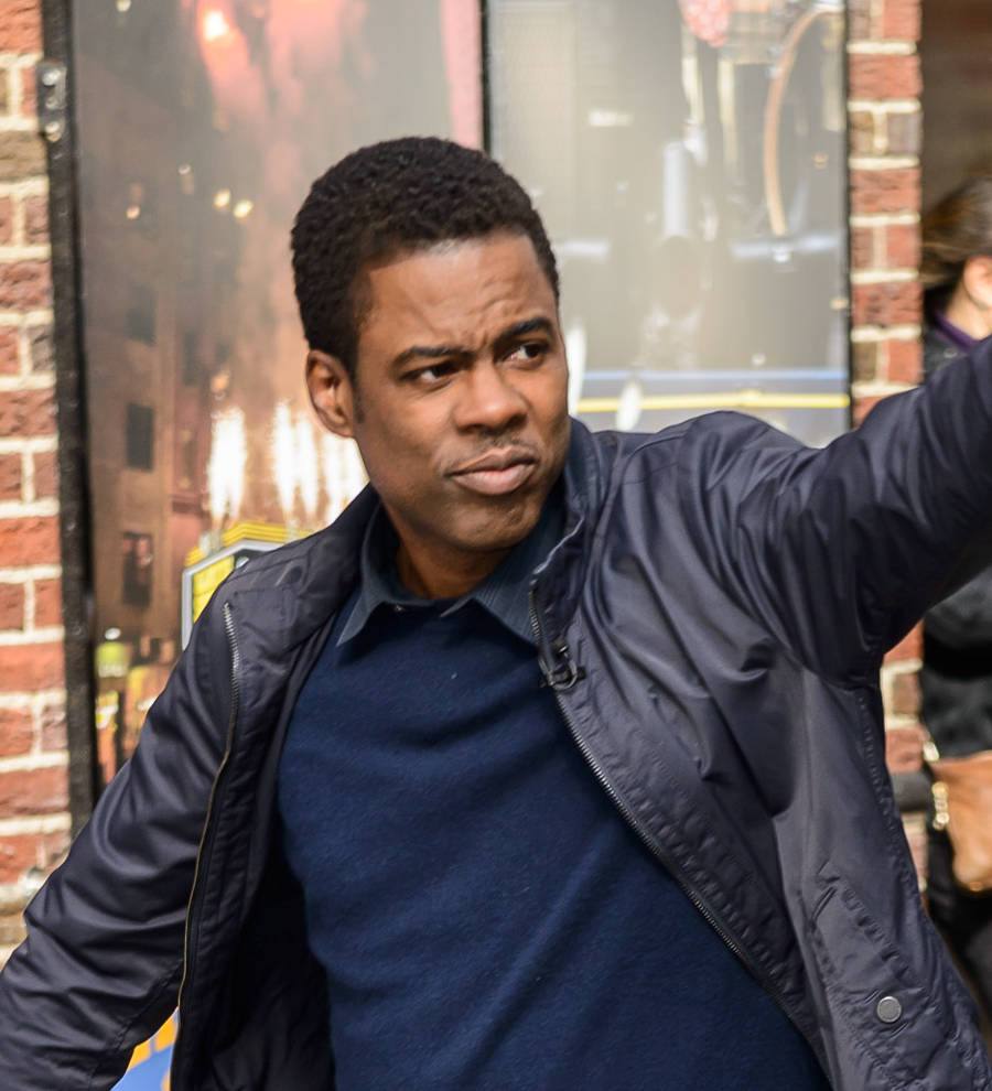 Chris Rock on stage in a dark suit, speaking into a microphone with a spotlight behind him