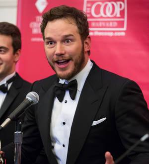 Chris Pratt smiles, waving behind a microphone at a press event