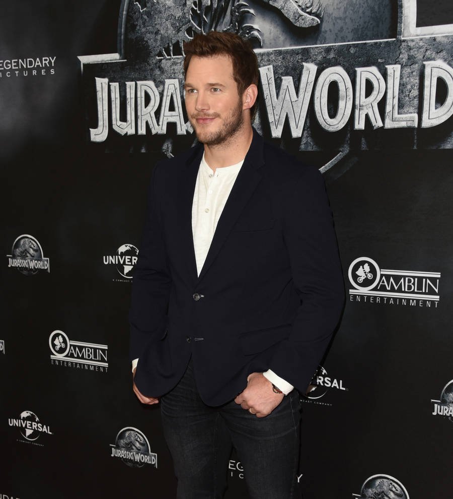 Chris Pratt stands on a football field in a blue jacket, smiling at the camera