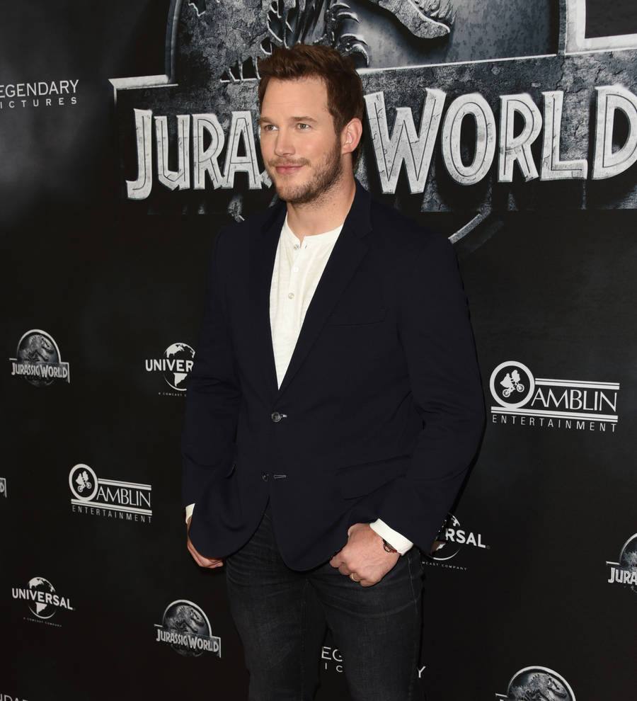 Chris Pratt stands on a football field in a blue jacket, smiling at the camera