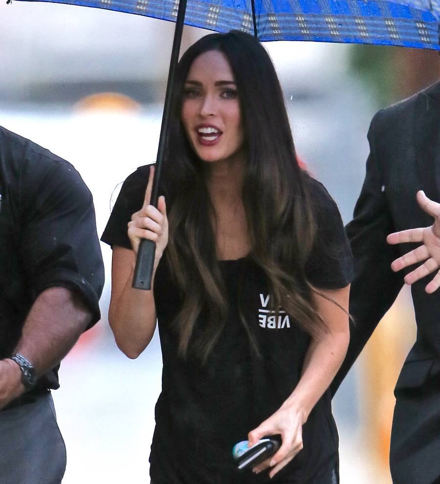 Megan Fox with long dark hair, wearing a white sleeveless top, smiling at the camera. Her hand touches her chest
