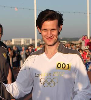 Matt Smith running in an Olympic torch relay, wearing a white tracksuit and holding a torch