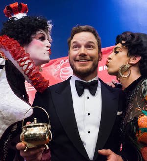 Chris Pratt, wearing a gold jacket and tiara, smiles with two Harvard students in a theatrical setting