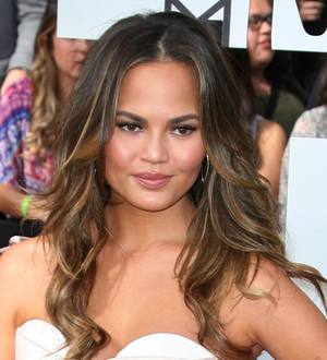 Chrissy Teigen poses in a black dress in front of a colorful, blurry background