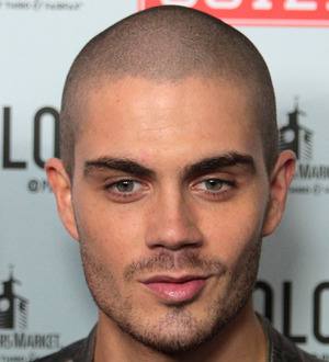 Max George smiling, wearing a dark jacket and white t-shirt, standing outdoors