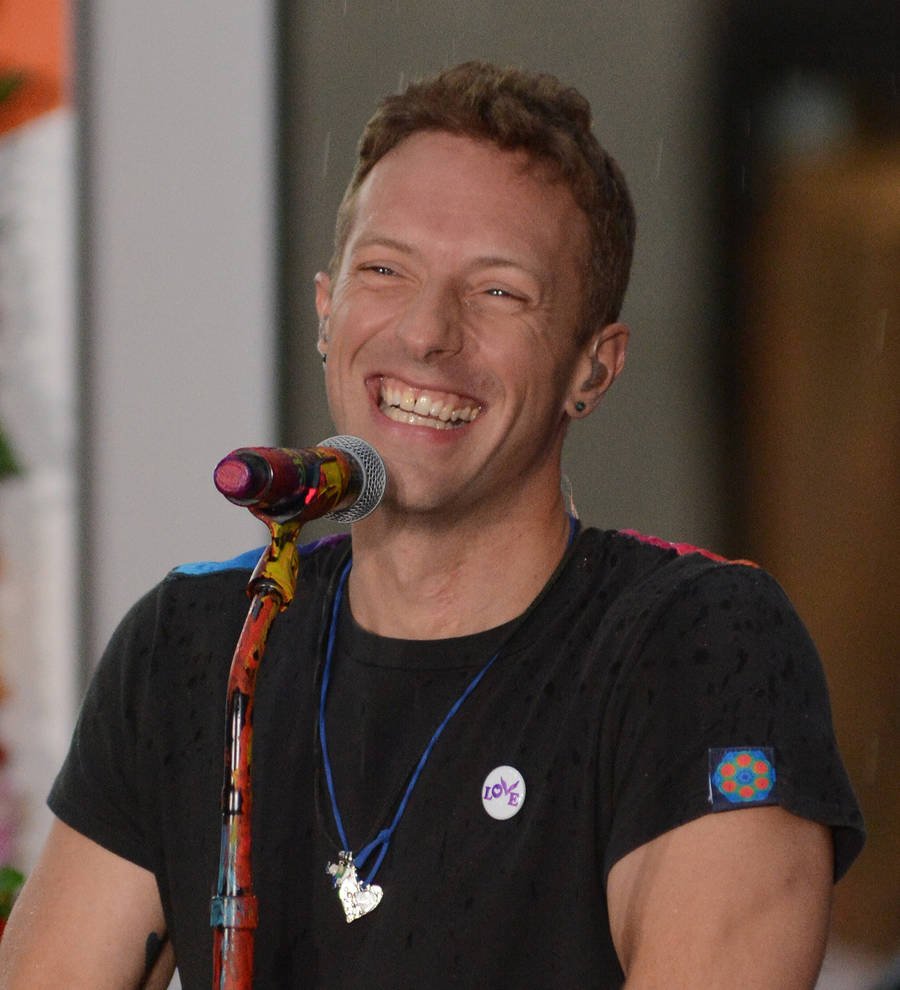 Chris Martin in a black shirt, seated at a table, signing papers, as a pen hovers above