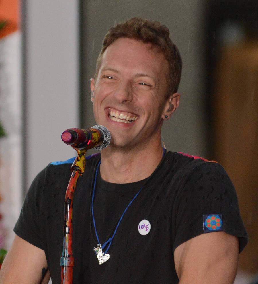 Chris Martin in a black shirt, seated at a table, signing papers, as a pen hovers above