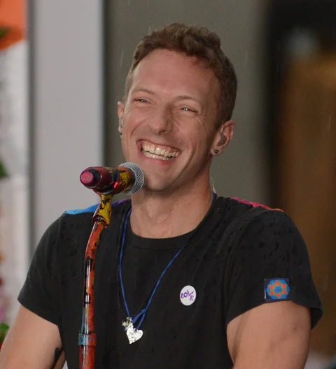 Chris Martin in a black shirt, seated at a table, signing papers, as a pen hovers above