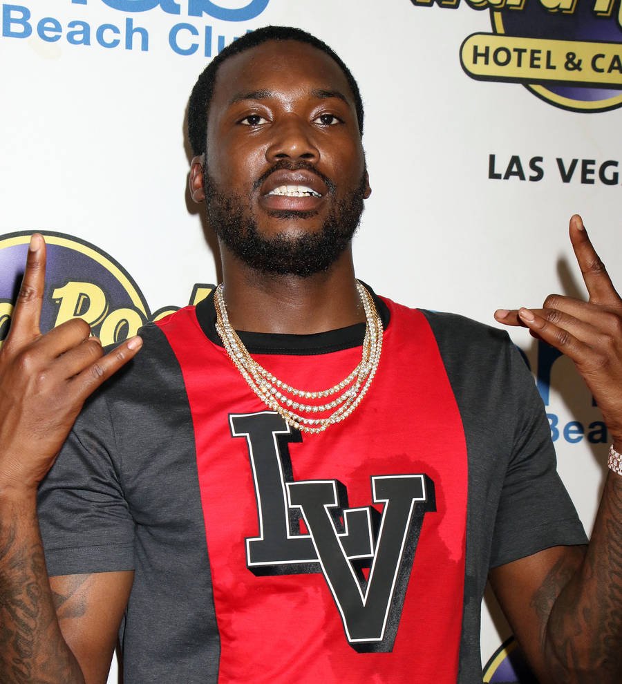Meek Mill stands in a hallway in a black and white striped shirt, looking seriously at the camera