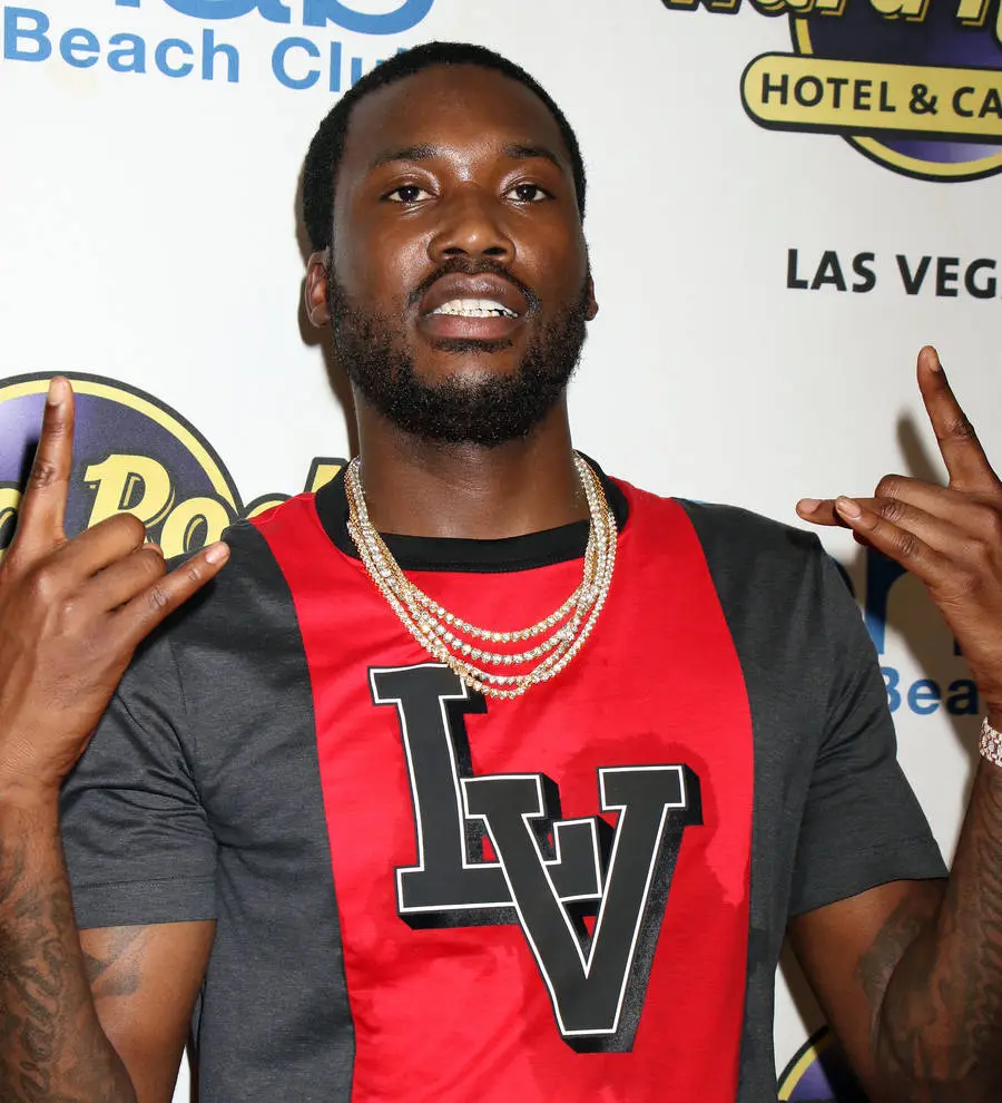 Meek Mill stands in a hallway in a black and white striped shirt, looking seriously at the camera