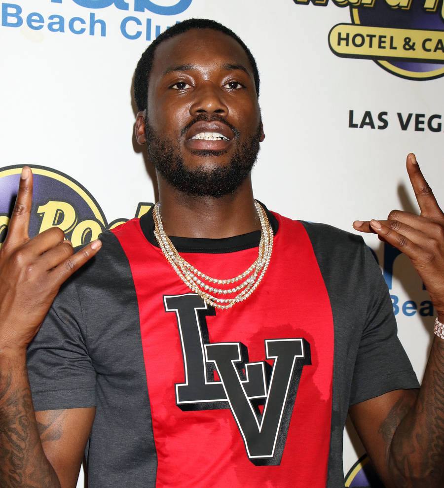 Meek Mill stands in a hallway in a black and white striped shirt, looking seriously at the camera